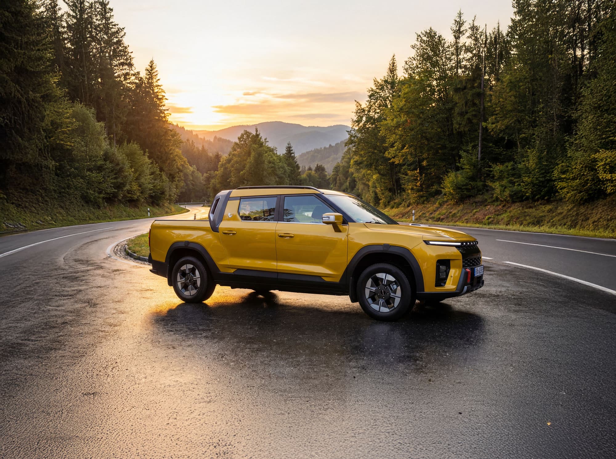 Yellow KGM Musso EV parked on a winding wet road, surrounded by lush green trees, with a sunset over distant hills in the background.