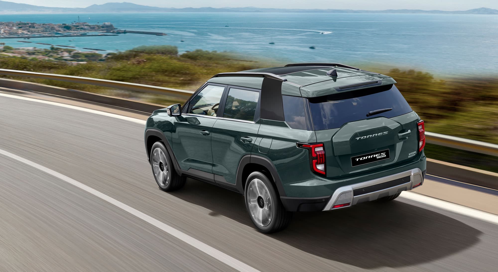 Green KGM Torres Hybrid driving on a coastal highway with a scenic view of the ocean and distant islands under a clear blue sky.