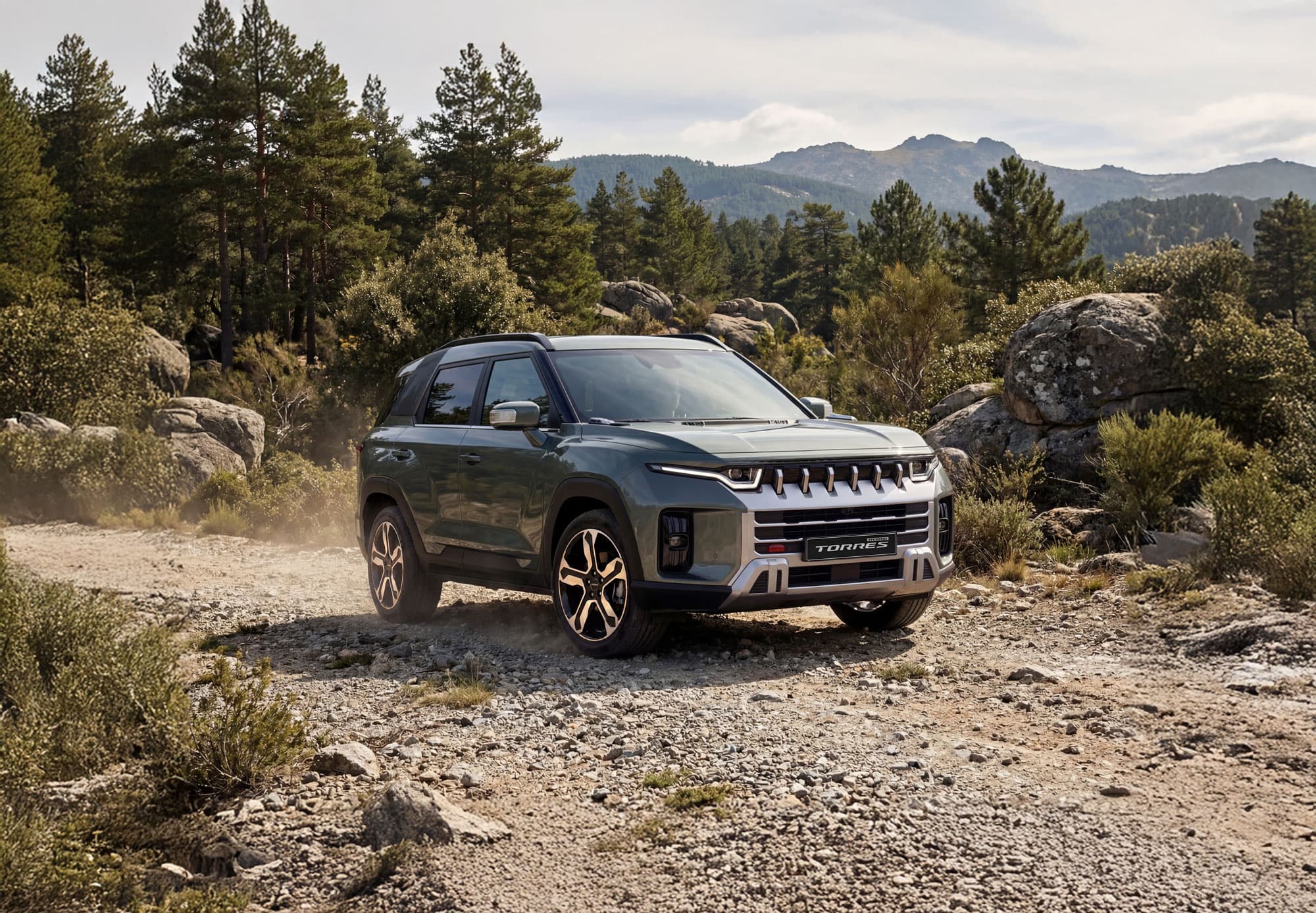 A rugged KGM Torres Hybrid driving on a rocky dirt path in a forested mountainous area, surrounded by trees and rocks under a clear sky.