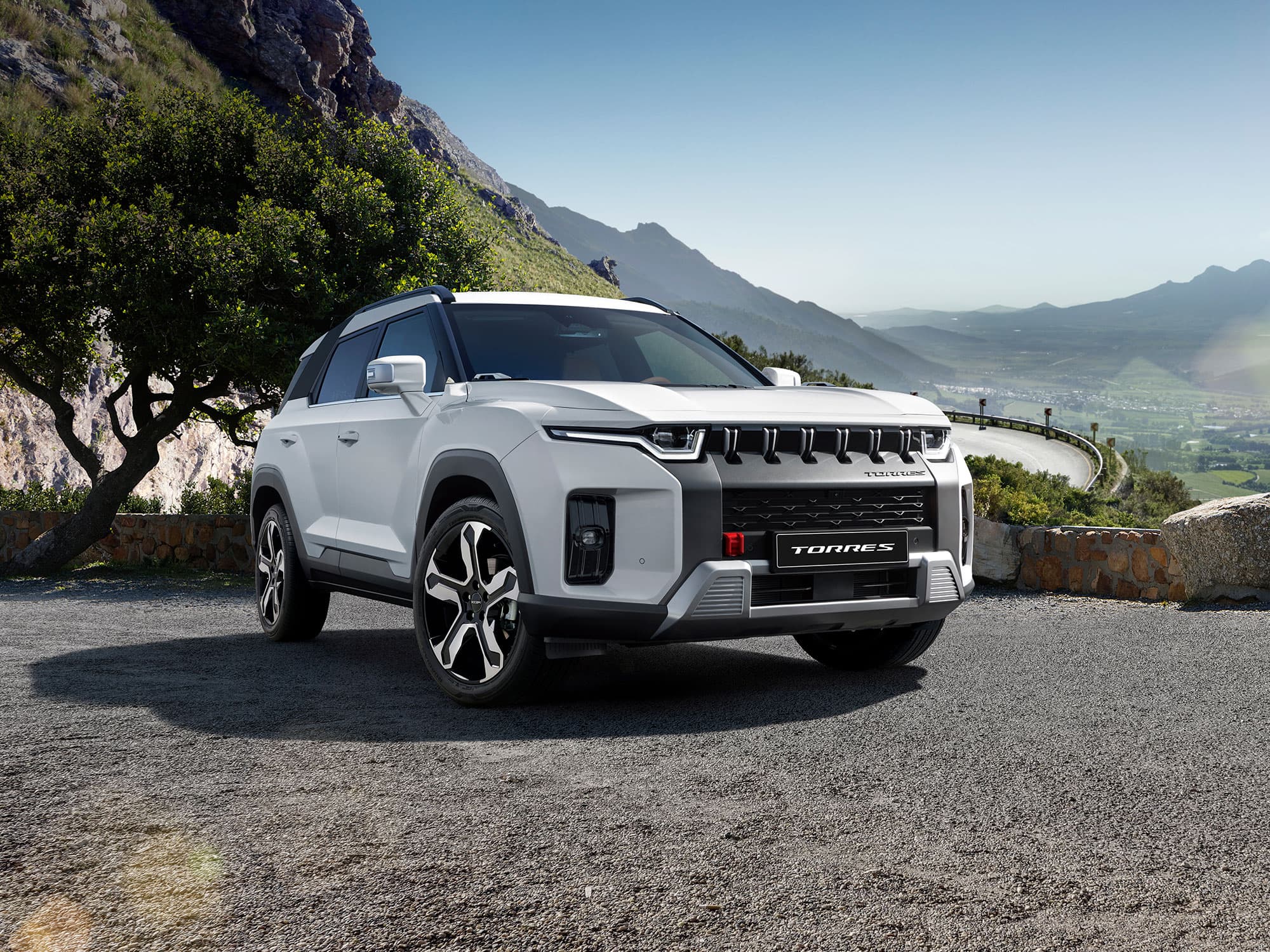A white KGM Torres Hybrid parked on a scenic mountain road with a winding path, overlooking a valley under a clear blue sky.