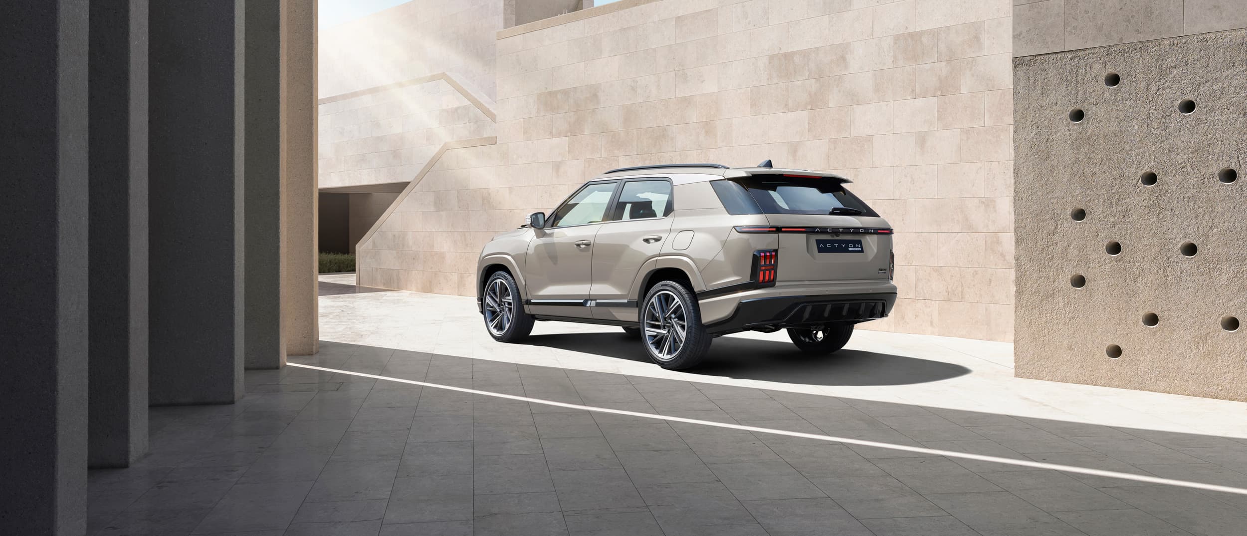 Beige KGM Actyon parked in a minimalist sunlit stone courtyard beside geometric walls and columns, casting a long shadow.