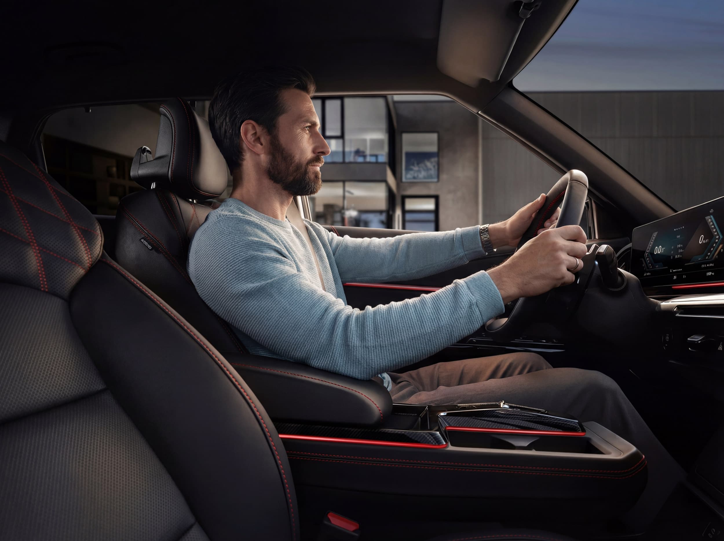 A man in a light blue sweater drives a modern car with black and red interior, focusing intently on the road ahead.